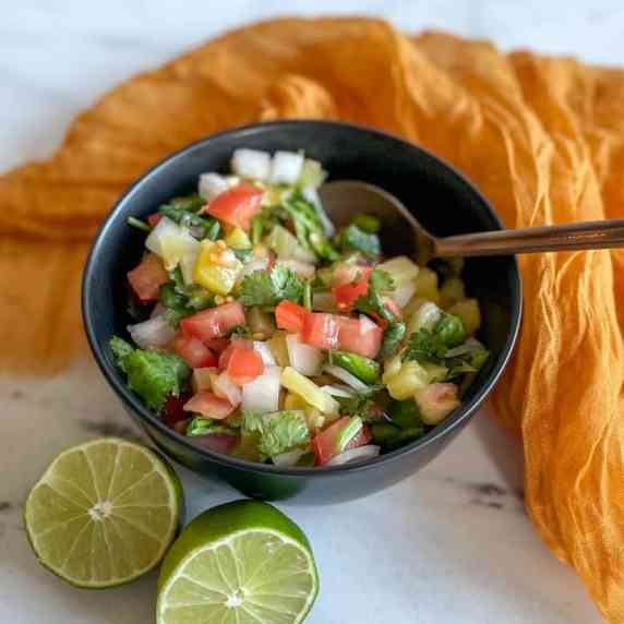 A bowl of pineapple pico de gallo is shown with a halved lime and a yellow linen.