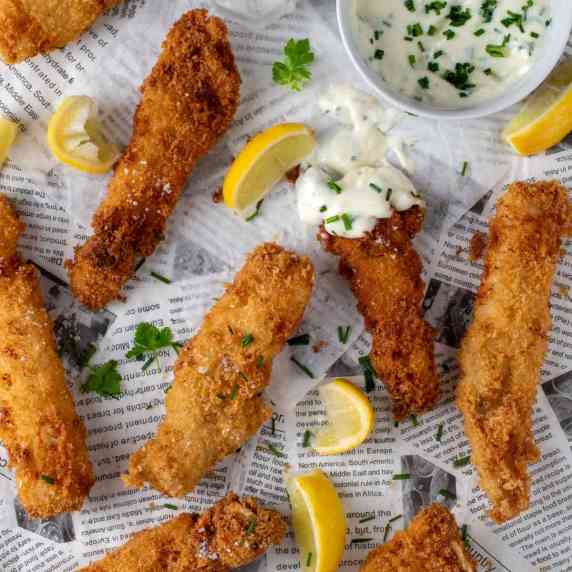 Fish goujons spread out on newspaper with some lemon slices and a garlic mayo dipping sauce on a sma