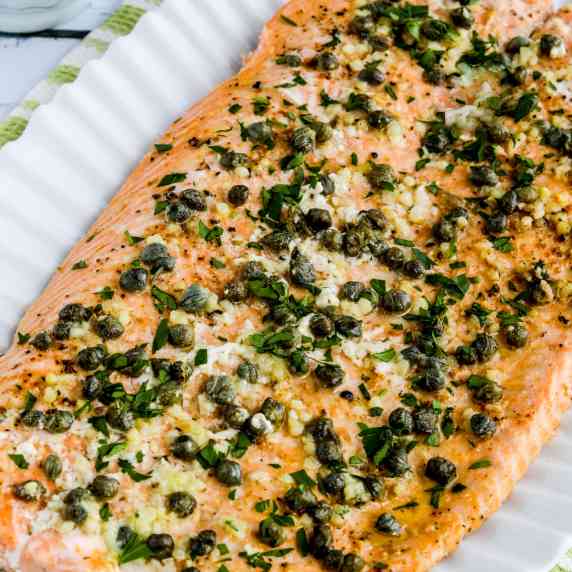 large salmon fillet on serving plate with butter, garlic, capers, and lemon