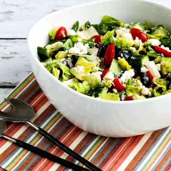 American Greek Salad with Lettuce shown in serving bowl with spoon, fork, and striped napkin.