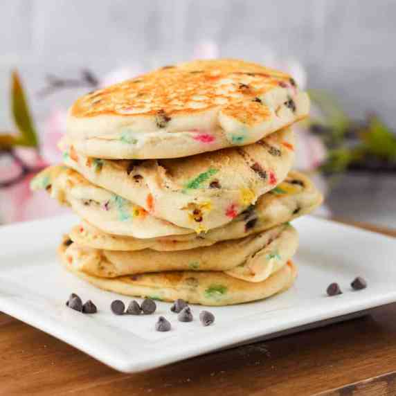 a stack of pancakes with sprinkles on a white square plate. the plate has chocolate chips on it.