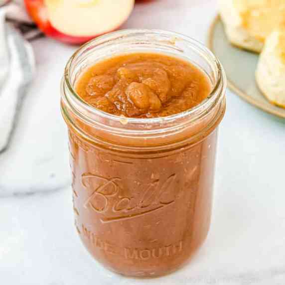 A mason jar of apple butter with apples and biscuits in the background.