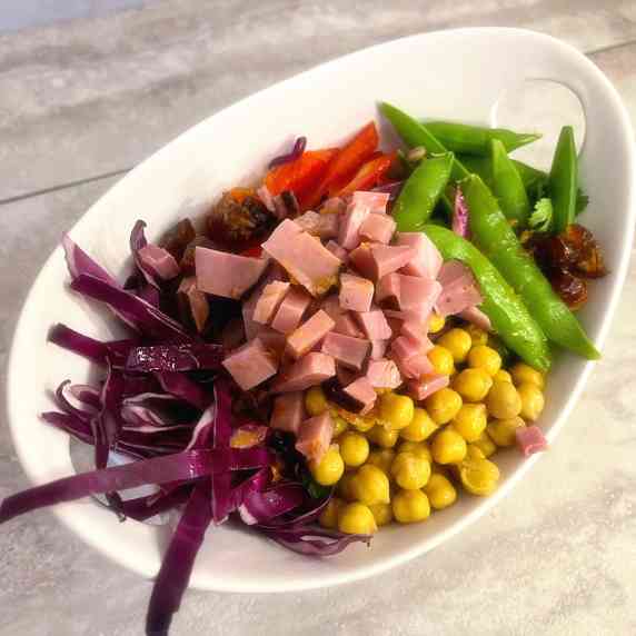 Barley, chickpeas, snap peas, red cabbage and dates arranged in a white bowl on a white countertop.