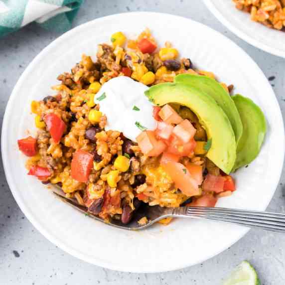 Ground beef burrito bowl covered in sour cream, avocado, and diced tomato with a fork in the bowl.