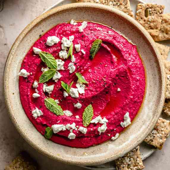 A bowl of beetroot hummus with olive oil, feta crumbs and mint leaves on top surrounded in crackers.