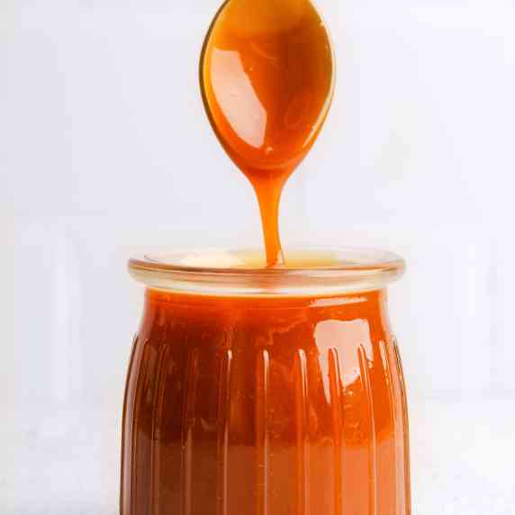 Homemade caramel sauce in glass jar with spoon on a white countertop