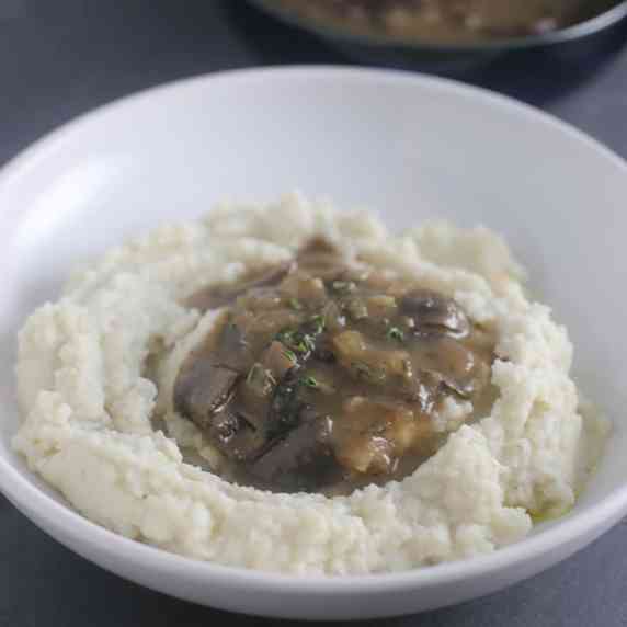 Mashed cauliflower topped with herby mushroom gravy in a white bowl. 