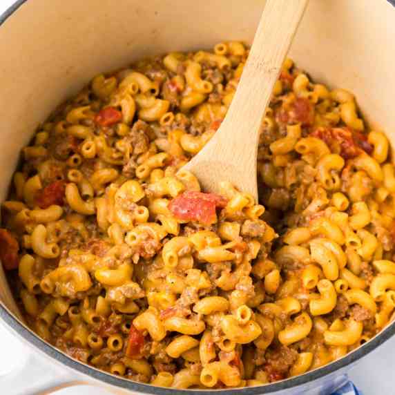 Inside a pot full of cheeseburger macaroni and pieces of tomato being stirred with a wooden spoon.