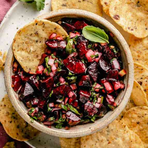 A bowl of cherry salsa on a plate with a round tortilla chip dipping into it.