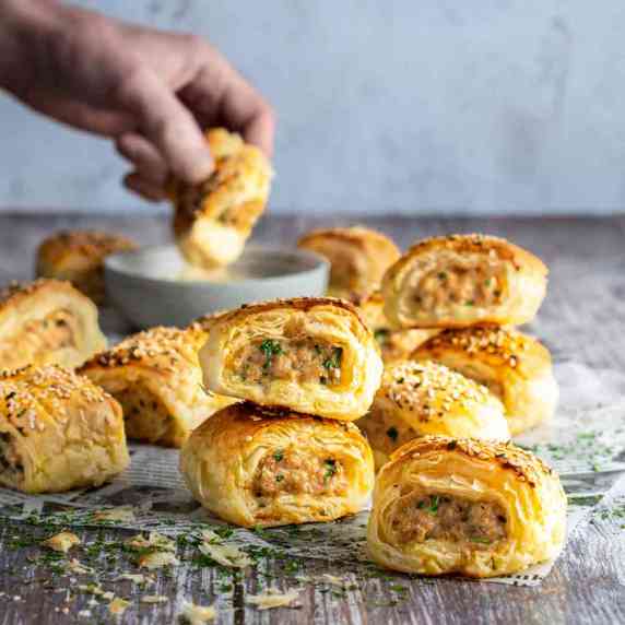 Chicken sausage rolls scattered on a table with a hand in the background dipping a sausage roll into