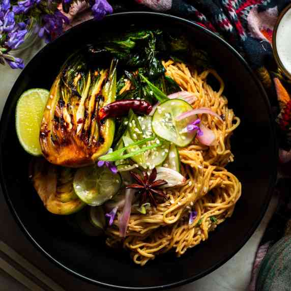 Noodles in a black bowl with grilled bok choy and pickled cucumbers on top.
