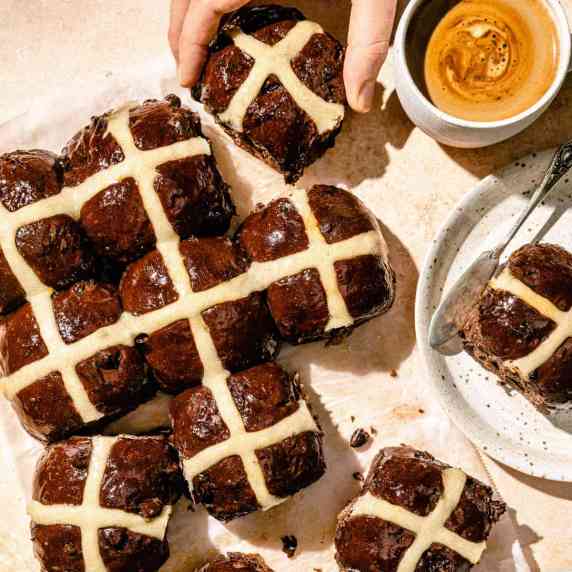 An array of chocolate hot cross buns on parchment with a plate and knife and coffee to the side.