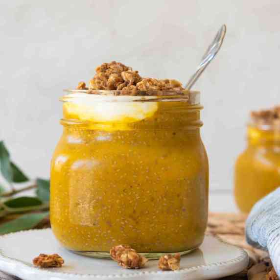 Two jars o pumpkin chia pudding on a beige plate with leaves and granola