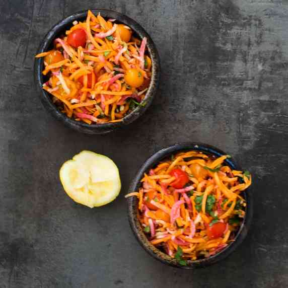 Grated carrot, ginger, and moo radish with halved cherry tomatoes and cilantro in black salad bowls.