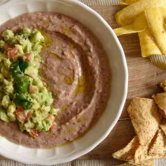 Red bean dip topped with avocado salsa on a serving board, with plantain and nacho chips