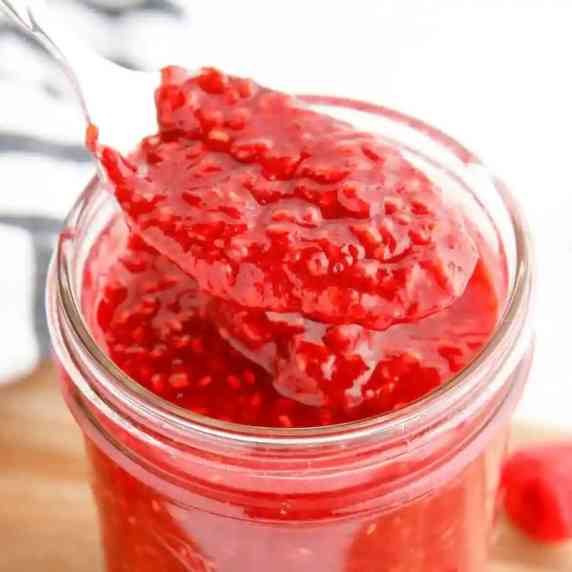 Close up side view of raspberry jam being lifted from a jar with a spoon cropped square.