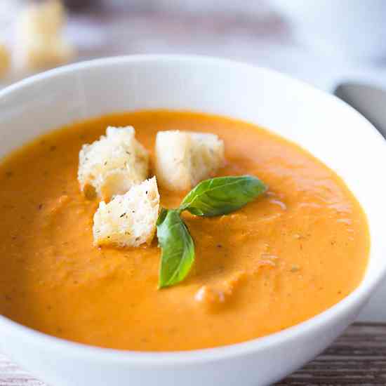 A white bowl filled with creamy tomato soup topped with three croutons and fresh basil.