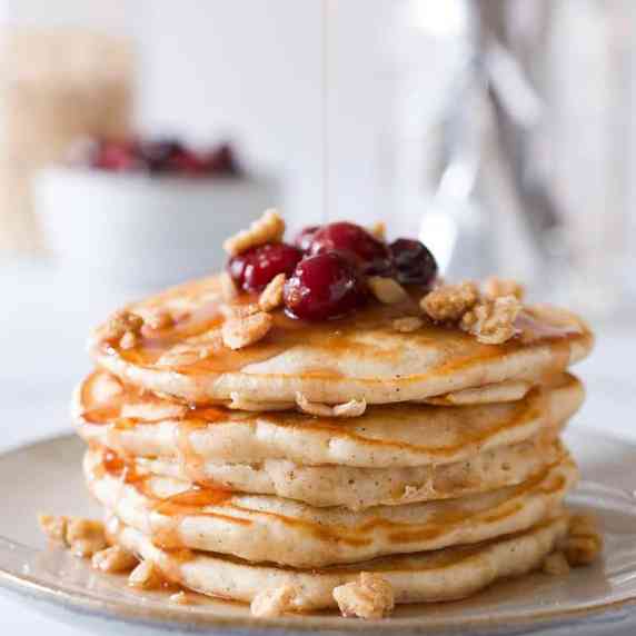 A stack of pancakes topped with cranberries and crumble being drizzled with syrup.