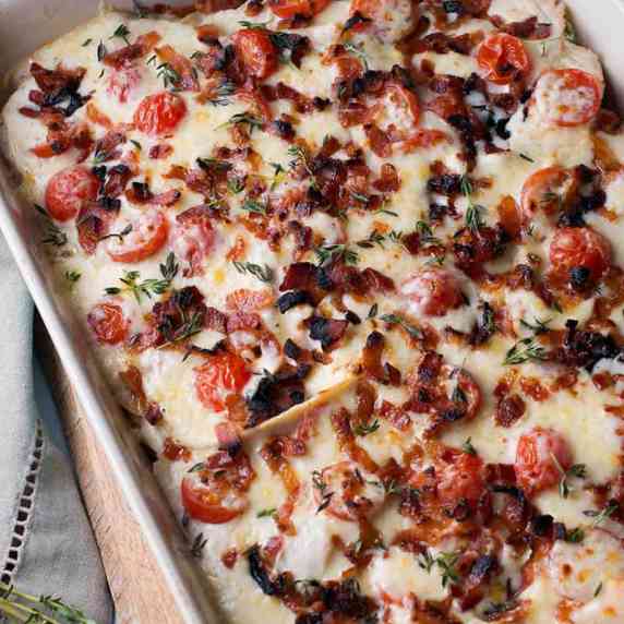 A casserole topped with gooey cheese sauce, cherry tomatoes, bacon bits and fresh thyme.