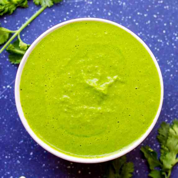 A limey green sauce in a white bowl on a blue countertop with cilantro around the bowl.