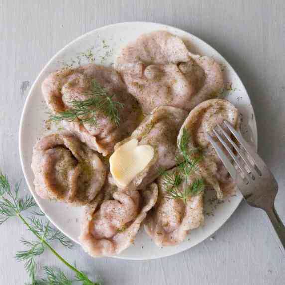 Pelmeni dumplings on a white plate with fresh dill on top, a piece of melting butter, and a fork.