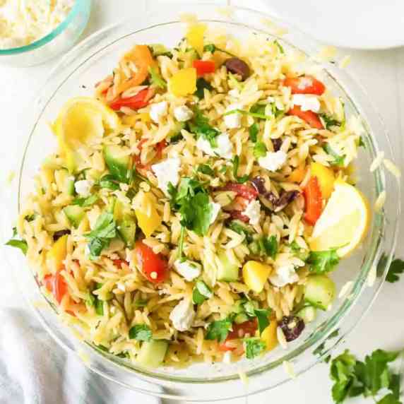 Mixed Greek Orzo salad with a wedge of lemon and feta cheese in a large bowl close up from overhead.