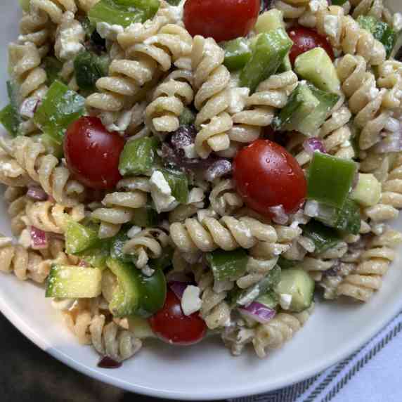 Pasta, green pepper, cucumber, tomatoes, red onion and feta cheese tossed and served in white bowl.
