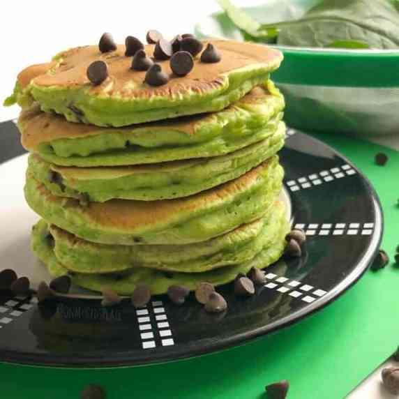 A stack of green spinach pancakes sits on a plate with a sprinkle of chocolate chips on top.