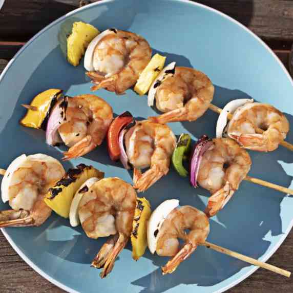A blue plate with three wooden skewers on it with shrimp, garlic, pineapple, onions, and pepper.