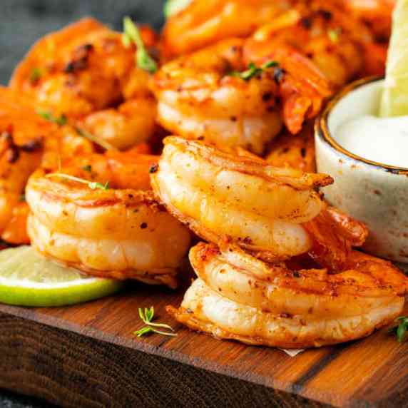 Wooden board with a pile of grilled chili lime shrimp and a creamy lime crema dipping sauce.
