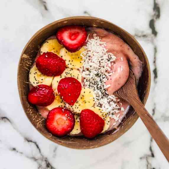 A spoon in the strawberry banana smoothie bowl that is topped with strawberries, banana, coconut.