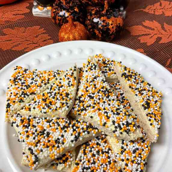 A white plate of Fairy bread with white, black and orange sprinkles, pumpkin ornaments in background