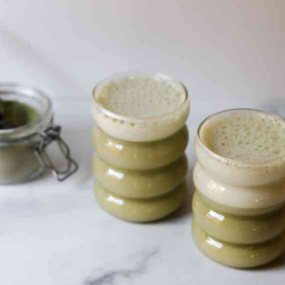 Matcha drinks in 2 glasses with matcha powder on the side all on countertop