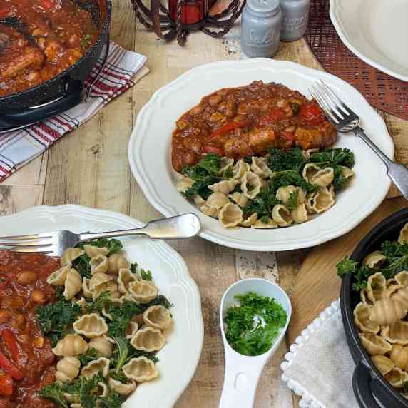 Two bowls of vegan sausage pasta with kale and pasta, serving dishes to side, and herbs,.
