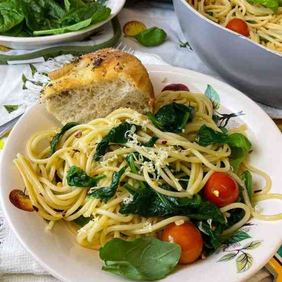 A bowl of garlic spaghetti with spinach and tomatoes, Italian bread and serving dishes.