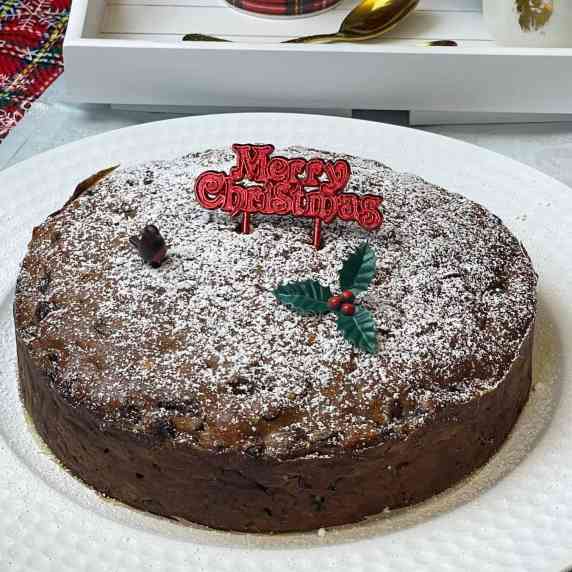 A Christmas cake sitting on a white serving plate, with little decorations on top, tea tray to side.
