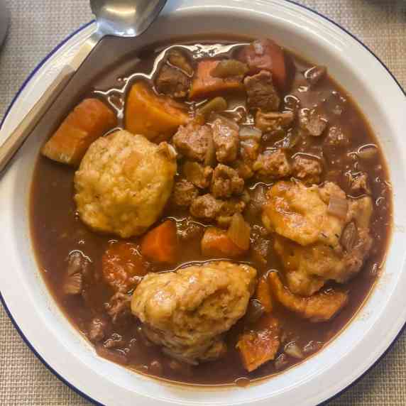 vegan beef stew, carrots, potatoes, soya chunks, and dark gravy, with dumplings, in a white bowl.