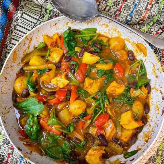A close up of vegan jalfrezi curry in a cooking pan, with a colourful patterned table cloth.