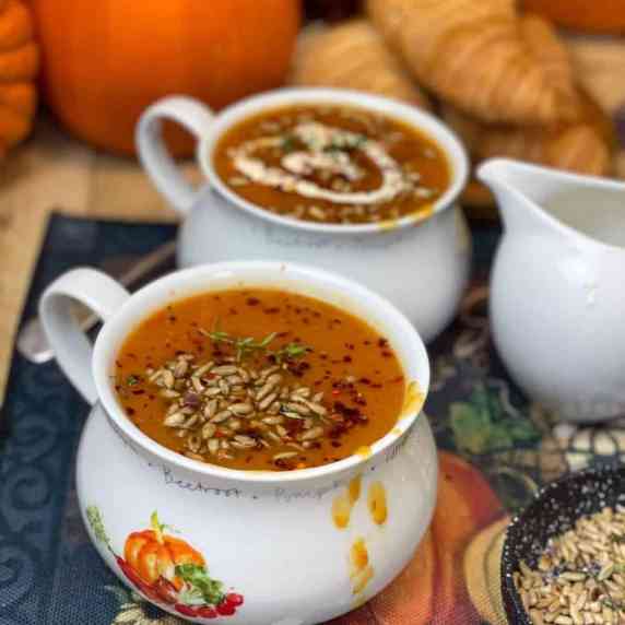 Two mugs of pumpkin soup with sunflower seeds and pumpkins in background.