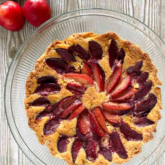 A round cake topped with a circular design in plums sits on a glass plate on a wooden surface.