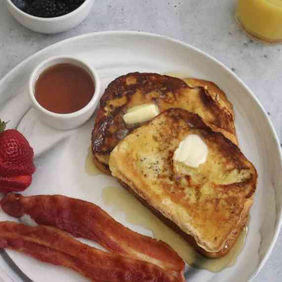 Two pieces of french toast topped with butter and maple syrup and served with bacon and strawberries