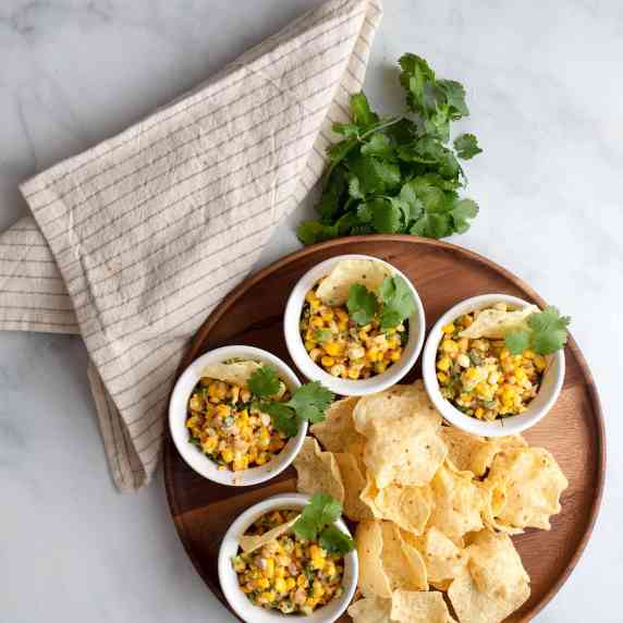 Four bowls of Mexican Street Corn accompanied by tortilla chips and cilantro