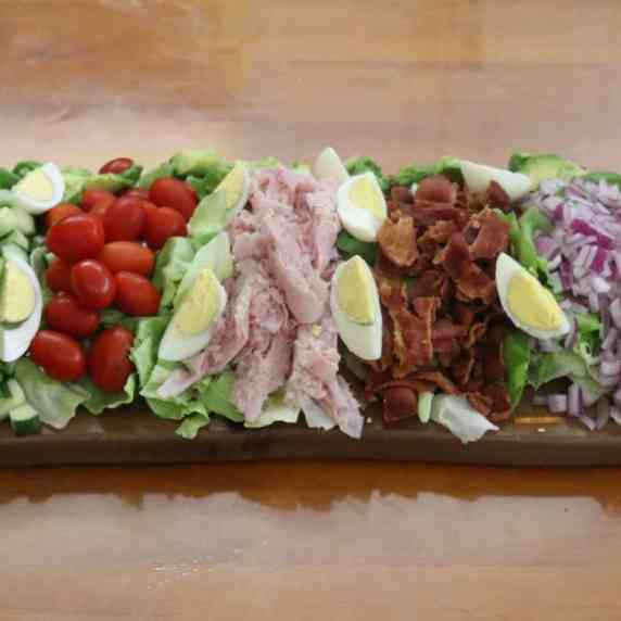Chef's salad with assorted vegetables and proteins layered in columns on a wooden board.