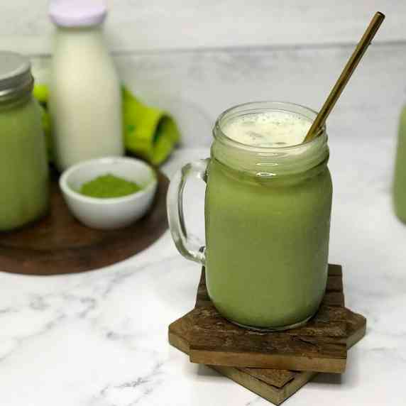 A vibrant green iced matcha latte in a mason jar glass with a gold straw sits on two wooden coasters