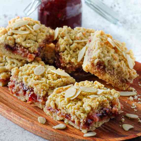 Six strawberry jam squares on a wooden board with a jar of strawberry jam on the background.