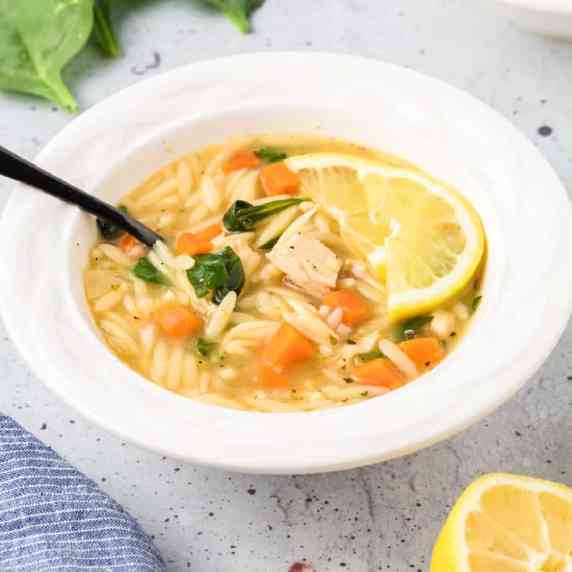Bowl of soup from the side with chicken, orzo, spinach, carrot and slices of lemon and a spoon. 