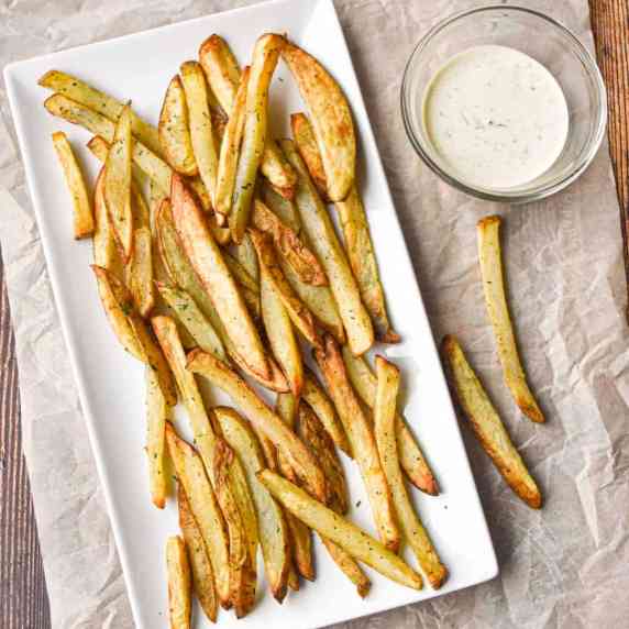 french fries on a white, rectangular plate next to a small bowl of ranch dressing on parchment paper