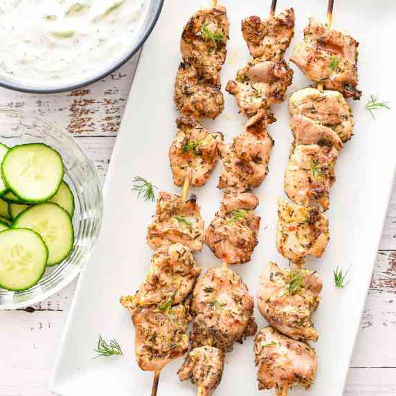 low fodmap chicken souvlaki on a white plate next to bowls of sliced cucumbers and tzatziki 