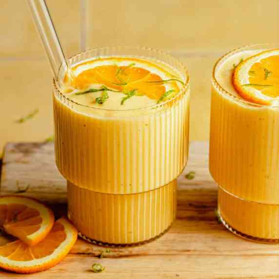 Two glasses of smoothie on a wooden board with orange slices on top and glass straws.