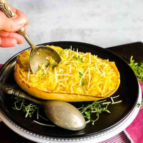 Cooked spaghetti squash on a black plate with a hand reaching in with a serving spoon.